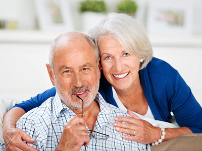 Ein lächelndes älteres Paar sitzt zusammen, der Mann hält seine Brille und die Frau umarmt ihn.